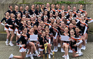 A large group of people dressed in matching cheerleading uniforms pose outdoors with trophies and signs, smiling and kneeling or standing on a paved surface in front of a wooden fence. Beschreibung von KI generiert