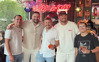 Five adults stand closely together indoors under a neon sign, smiling and holding drinks, with one person wearing a black graphic t-shirt and glasses. Beschreibung von KI generiert