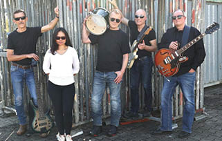Five adults wearing casual clothes and sunglasses stand outdoors on a paved surface with musical instruments including guitars and a drum. Beschreibung von KI generiert