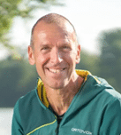 A smiling person wearing a green jacket with a yellow inner lining is outdoors with a blurred natural background. Beschreibung von KI generiert