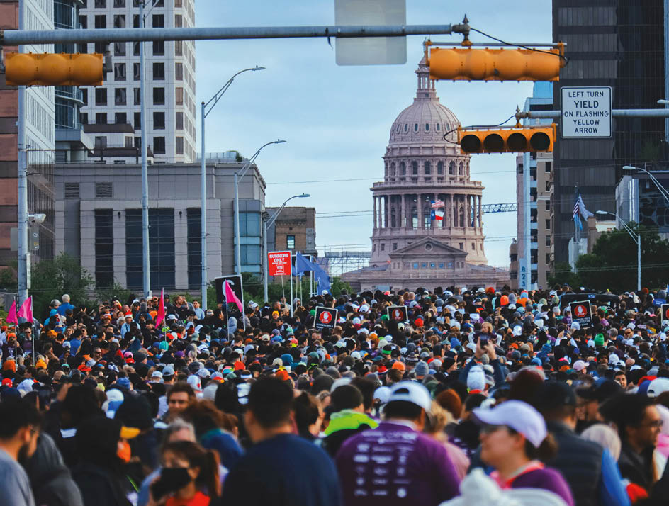 A large group of people wearing athletic clothing are running in a race on a road lined with spectators and banners. Beschreibung von KI generiert