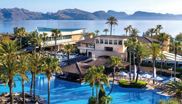 A luxurious resort with a large blue swimming pool surrounded by palm trees and lounge chairs overlooks a calm body of water with mountains in the background. Beschreibung von KI generiert