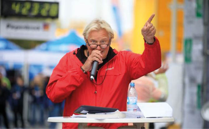 A person wearing glasses and a red jacket is speaking into a microphone while standing behind a table with papers and a water bottle, pointing upwards with one hand. Beschreibung von KI generiert