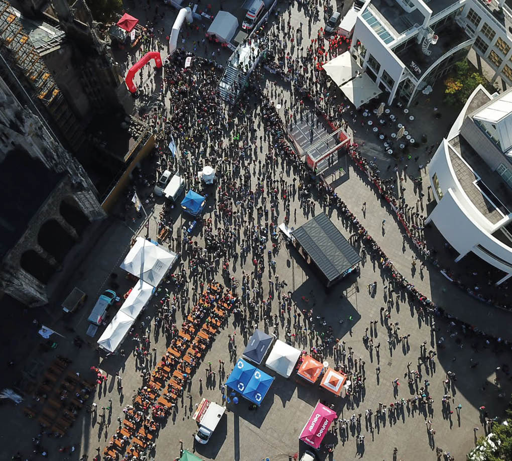 A large crowd of people gathers in an outdoor urban plaza with tents, tables, and booths arranged in rows, casting long shadows in the sunlight. Beschreibung von KI generiert