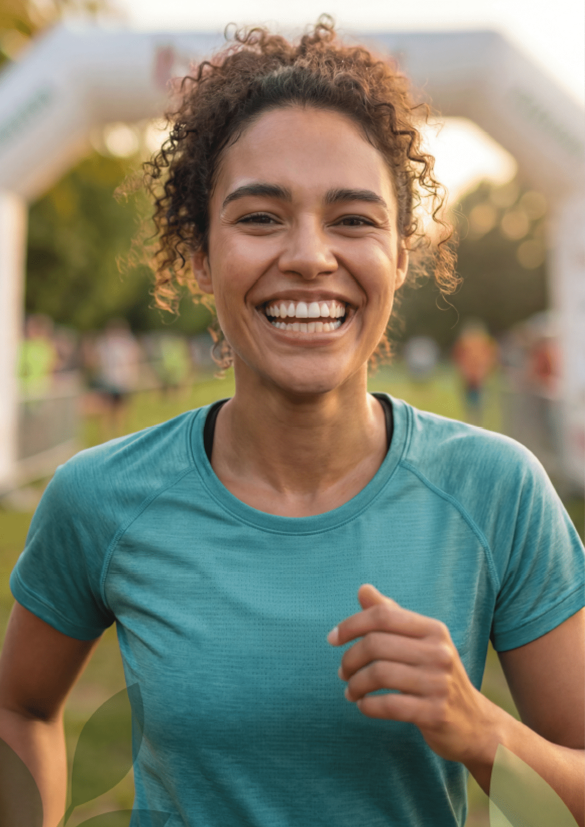 A person with curly hair wearing a teal shirt is smiling broadly while running outdoors near a race finish line. Beschreibung von KI generiert