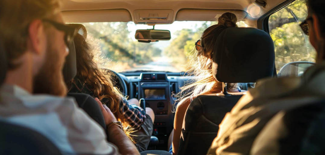 Four people with casual clothing and sunglasses are sitting inside a car during daylight, facing forward as the vehicle travels on a sunlit road surrounded by trees. Beschreibung von KI generiert