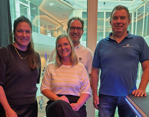 Four adults smiling and posing indoors near large windows with a modern architectural background. Beschreibung von KI generiert