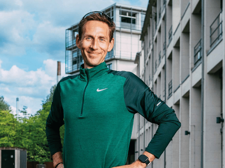 A person wearing a green and black Nike long-sleeve top and a smartwatch is standing outdoors with hands on hips, smiling in front of a modern building. Beschreibung von KI generiert