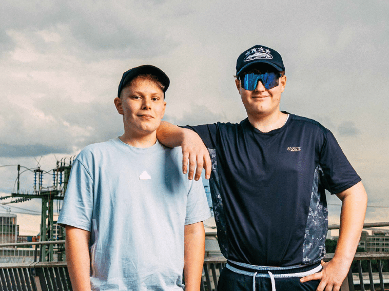 Two adults wearing casual clothing and caps stand close together outdoors with one resting an arm on the other's shoulder against a cloudy sky background. Beschreibung von KI generiert