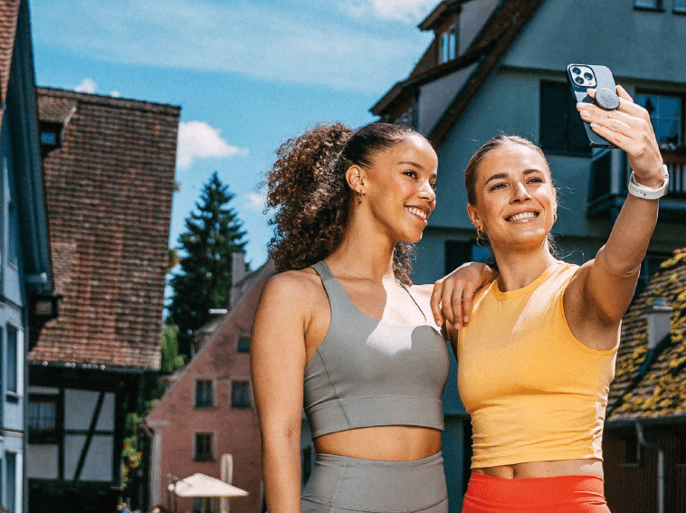Two adults wearing athletic clothing are smiling and taking a selfie outdoors in a village setting with traditional buildings in the background. Beschreibung von KI generiert