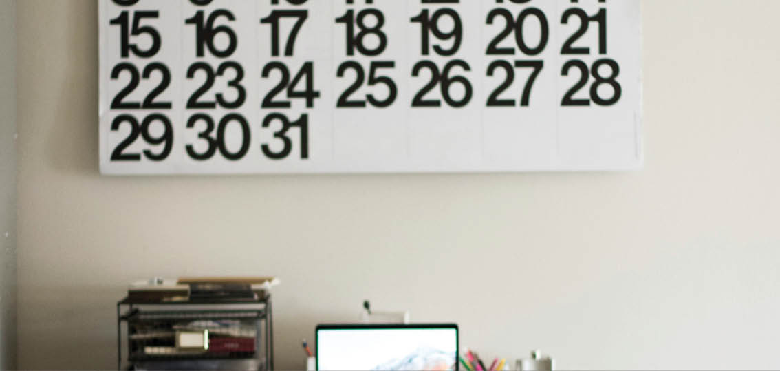 A workspace with a laptop on a desk, a calendar with large black numbers on the wall above, and various office supplies nearby. Beschreibung von KI generiert