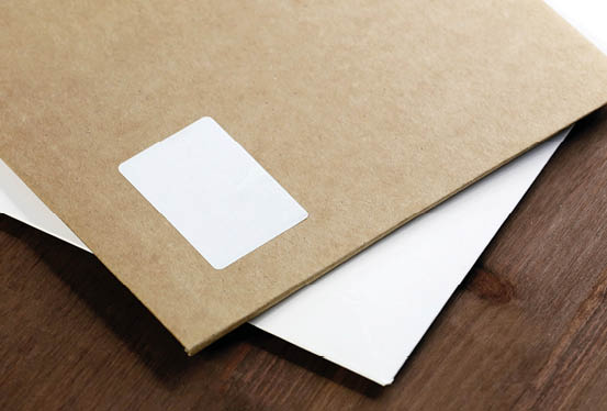 A brown envelope with a white label partially covering a white envelope is placed on a wooden surface. Beschreibung von KI generiert